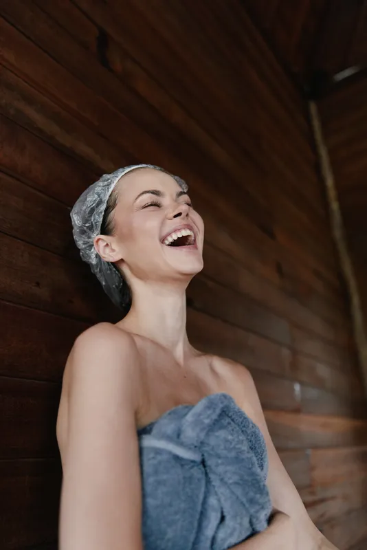 laughing woman wearing shower cap wrapped in towel enjoying spa moment against wooden wall indoors, natural light creating warm atmosphere and relaxation