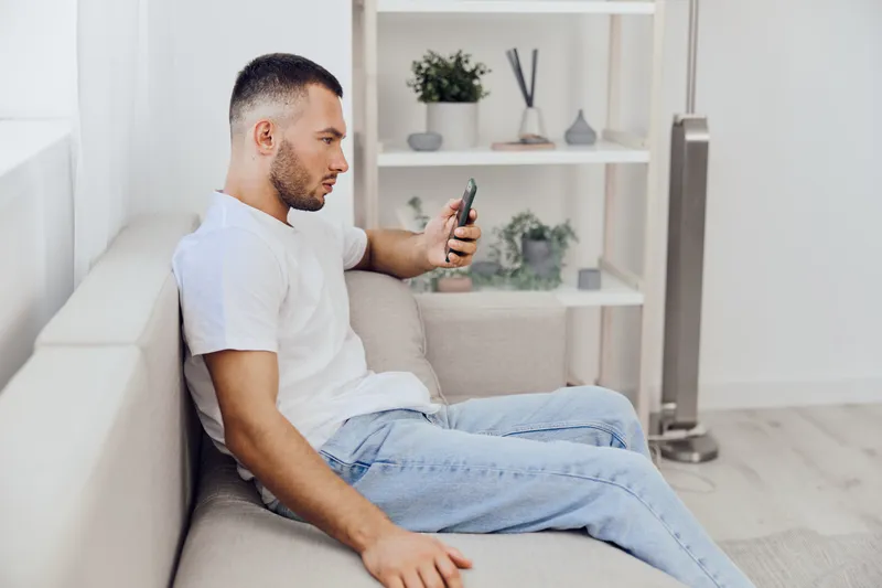 Young man focused on smartphone, sitting comfortably on a sofa in a modern, minimalistic room with light tones and greenery, reflecting contemporary lifestyle trends.