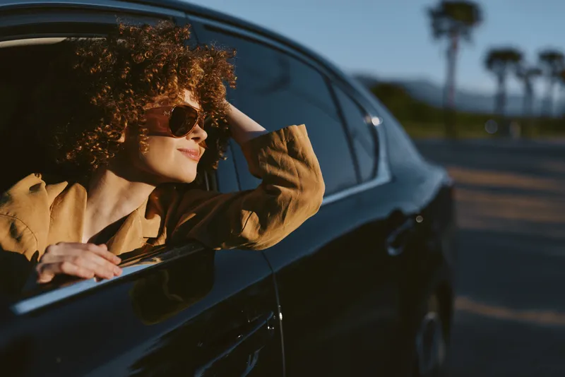 Woman leaning from a glossy car window, basking in sunlit breeze, casual lifestyle moment with retro color tones, relaxed pose, warm shadows, open road backdrop