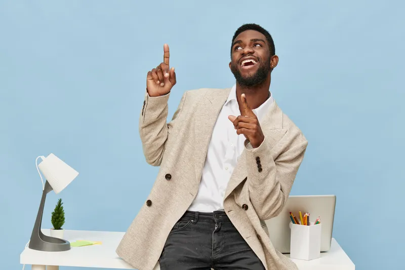 Confident young African American man in beige blazer and white shirt expressing joy, pointing upward with his finger while standing in modern office with desk, lamp, and plant, bright blue background,