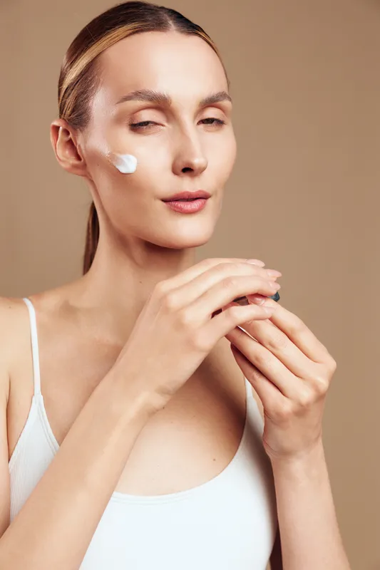 Young woman with blonde hair applies moisturizer on her cheek while holding a cream jar against a neutral background in a calm skincare routine moment.