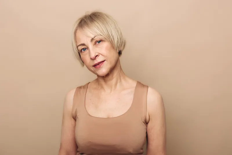 Portrait of a mature woman with short blonde hair wearing a beige tank top against a neutral background. Concept of natural beauty, aging gracefully, and confidence in simplicity.