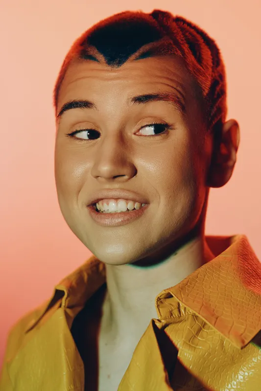 young woman with short braided hair wearing yellow textured jacket smiling and looking sideways against warm peach background, creative studio portrait