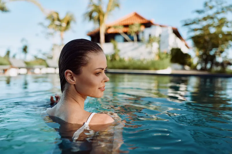 Young woman in white swimsuit relaxing in outdoor pool with tropical palm trees and villa in background. Concept of summer lifestyle, leisure, and vacation time.