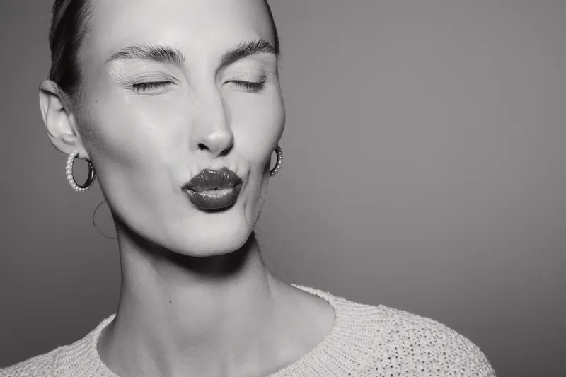Close-up of a young white woman with pale skin and earrings making a playful kiss face with closed eyes on grey background.