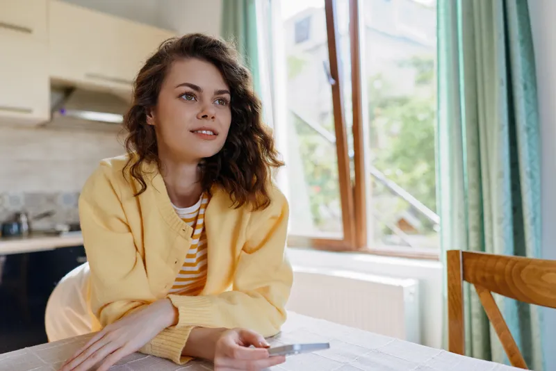 Curious woman with curly hair sits at a kitchen table in a yellow blazer gazing into the distance Natural daylight highlights a cozy home setting casual lifestyle and everyday creativity