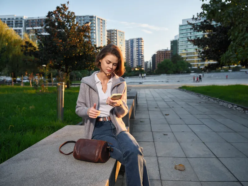 A focused young professional woman sits on a bench in a modern city park. She looks at her smartphone with concentration, creating a calm and contemporary urban moment.