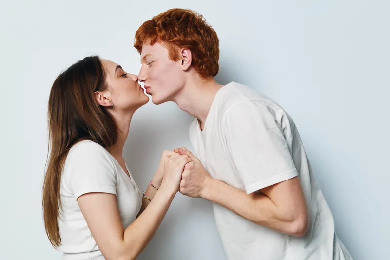 Young couple with casual clothes kissing and holding hands on light background. Romantic relationship, love, affection and intimacy between man and woman in studio.