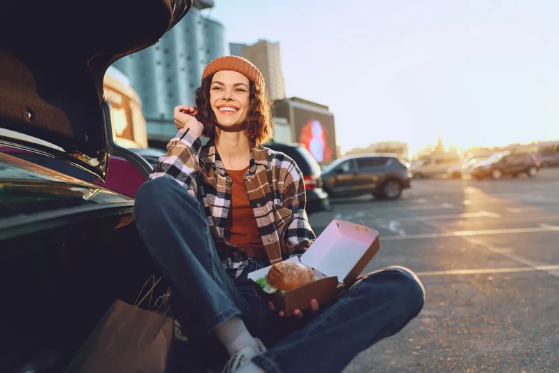 Woman sits at car trunk in parking lot eating takeout, smile and beanie, jeans and plaid shirt at sunset; candid lifestyle shot with authenticity, golden hour glow, mindful living, emotional