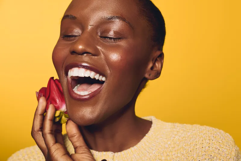 Happy African woman with dark skin laughs joyfully holding red rose near face. Yellow background and cozy sweater create warm and vibrant mood. Concept of beauty, happiness, and nature.