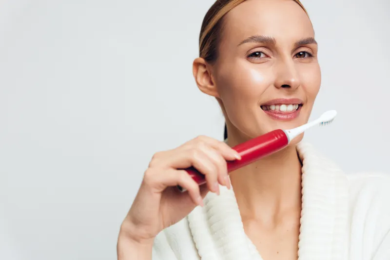 Smiling woman holding electric toothbrush, showing oral hygiene routine, clean teeth focus, bright background, casual white sweater, healthy lifestyle concept.