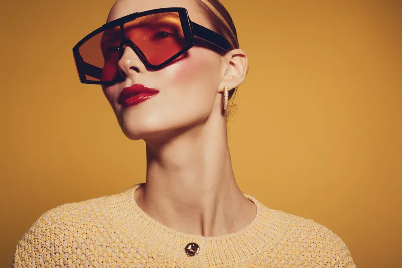 Stylish woman with short hair wearing oversized sunglasses and red lipstick poses against warm yellow background. Fashion portrait with modern accessories and confident expression.