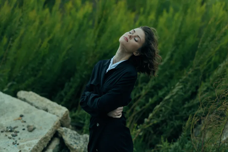 Woman in a dark coat leans back with closed eyes in an outdoor nature scene, a breezy pose that conveys fashion, elegance, serenity and calm mood amid lush greenery