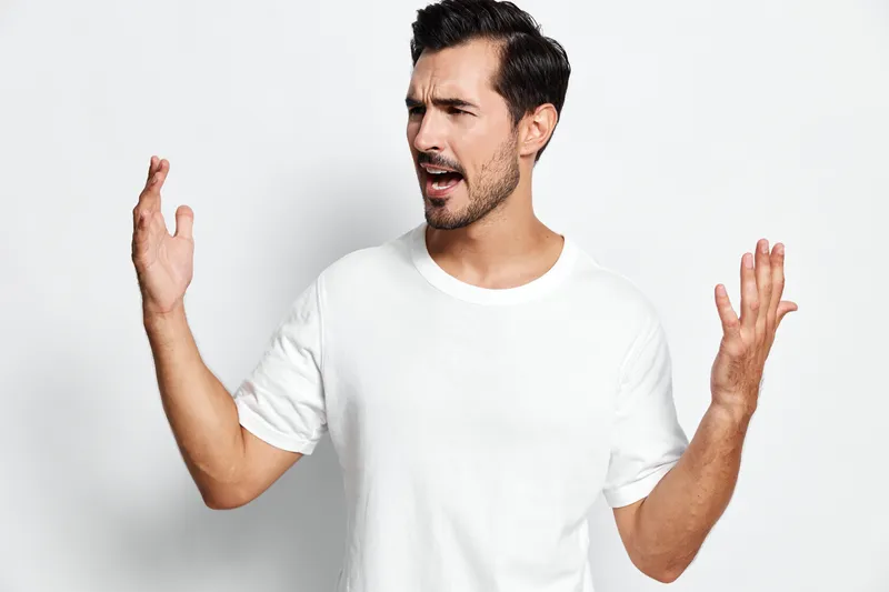 Expressive man gesturing with hands in frustration, wearing a casual white t-shirt, against a light grey background. Emotion and communication concept.