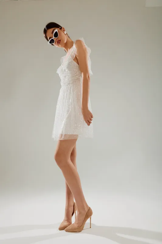Elegant woman in a white lace dress and high heels poses confidently wearing sunglasses. Studio shot on a neutral background. Concept of woman beauty fashion and style.