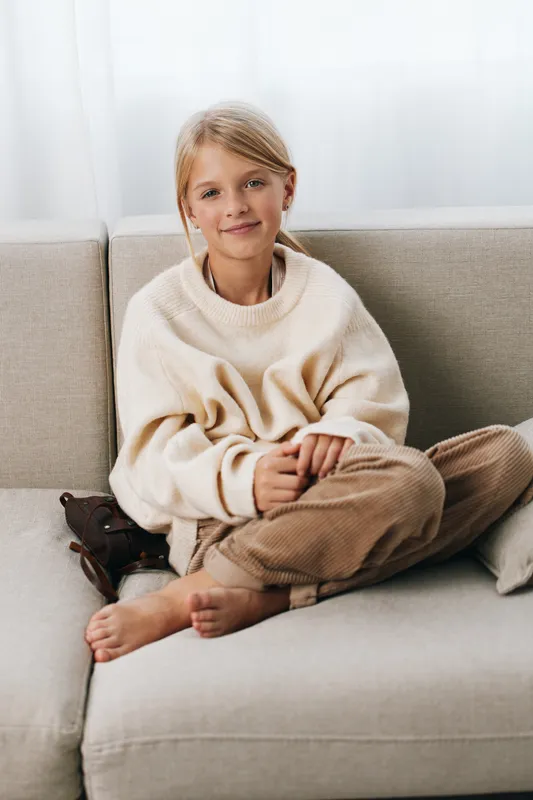 A young girl sits comfortably on a light sofa, wearing a cream oversized knit sweater and soft brown pants, creating a warm, relaxed portrait with soft natural light.