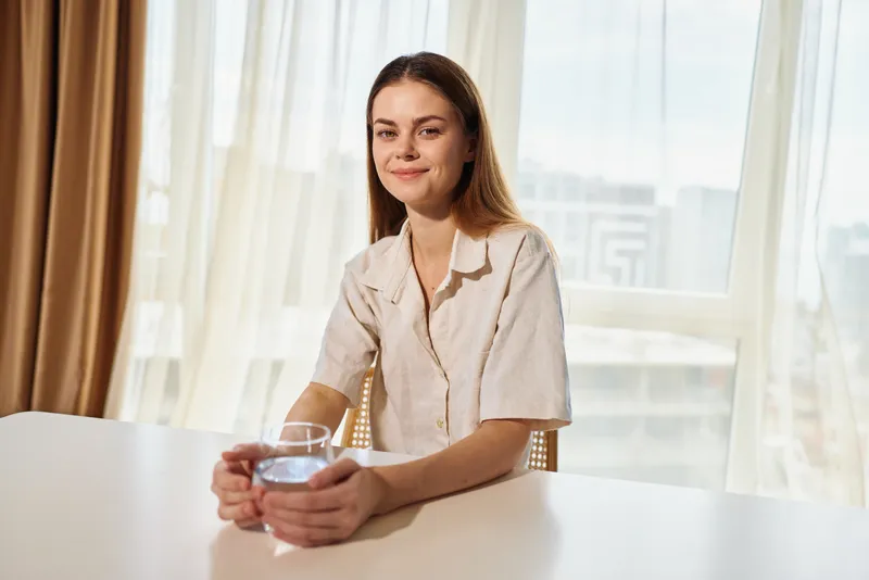 Woman smile holding glass of water at table in home portrait, morning light through window and curtains, casual female sitting relaxed, drinking for wellness, hydration and calm.