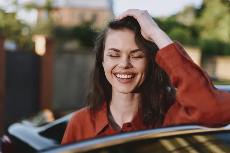 Lifestyle portrait of a cheerful woman in a red shirt, smiling with hand on hair, outdoor lighting by a car, casual candid moment for lifestyle storytelling