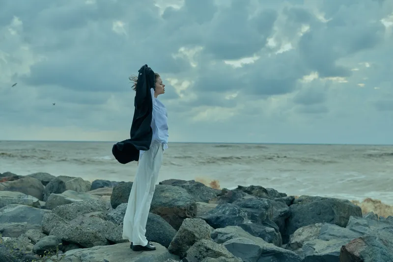 Woman standing on jagged rocks by the sea, outdoor scene, arms raised toward the windy sky, expressing freedom and stretch, coastal landscape, casual outfit, adventure and resilience against waves,
