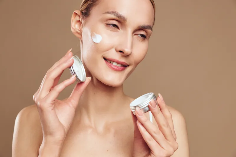 Smiling woman with clear skin applying white face cream on cheek, holding open jar. Close-up on beige background. Concept of skincare, beauty routine, wellness, self-care, hydration.