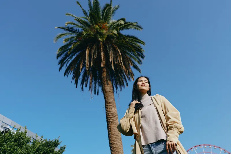 Woman outdoor palm tree against blue sky, showcasing fashion casual jacket and sunny stance in a bright daylight street scene with confident pose and relaxed mood.