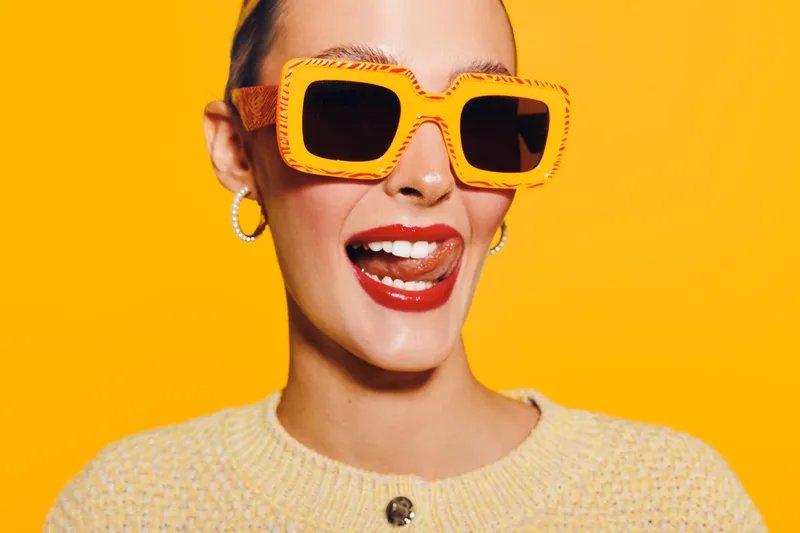 Young woman with orange sunglasses and red lipstick smiles playfully with tongue out on bright yellow background. Fashion portrait with trendy accessories and joyful expression.