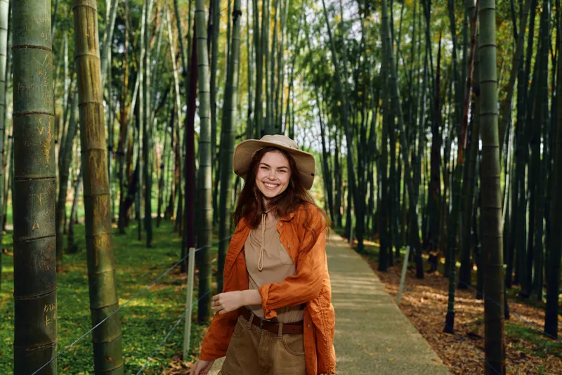 woman bamboo forest path smiling hat walking in a sunlit grove, casual traveler in straw hat and orange cardigan strolling along a paved trail among tall green stalks for nature escape