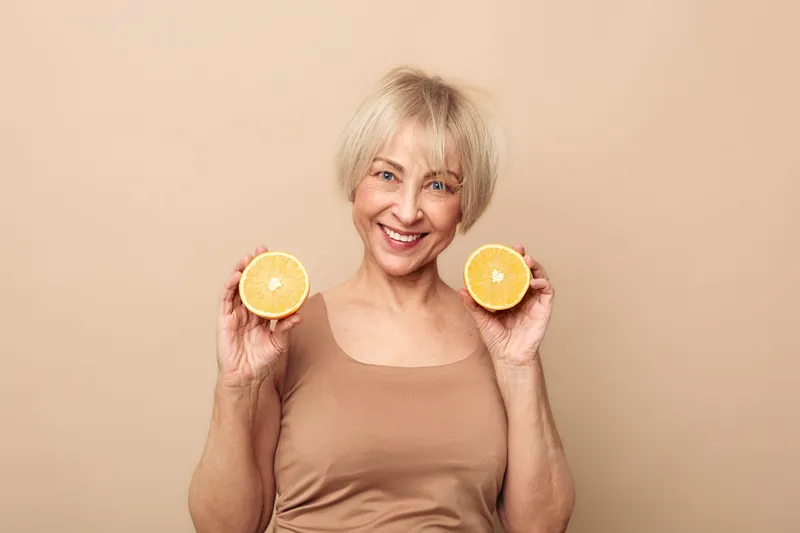 Smiling mature woman with short blonde hair holds two halves of a fresh orange close to her face, wearing a beige top against a soft beige background, concept of health and natural beauty.