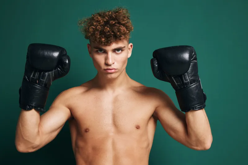Young male boxer with curly hair wearing black boxing gloves posing shirtless against dark background. Athlete showing strength and focus for training or competition.