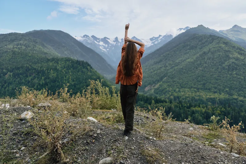 A lone hiker stands on a rocky overlook facing majestic mountains with snow caps, arms raised in triumph against a vast, tranquil landscape.
