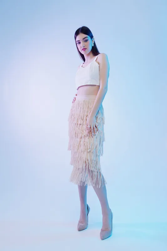 Young woman in a white crop top and fringed skirt poses confidently in high heels on a gradient background. Concept of fashion, beauty, and style.