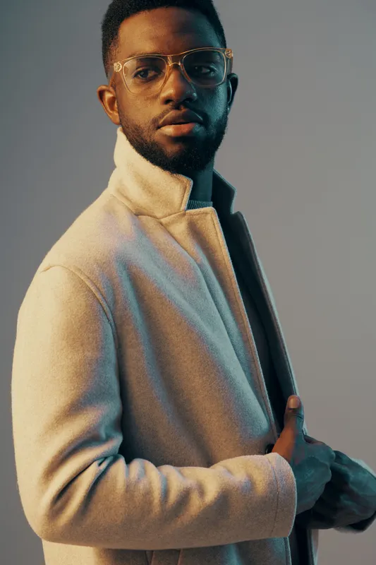 Stylish black man in modern attire with glasses posing confidently against a colorful backdrop, embodying sophistication and contemporary fashion trends