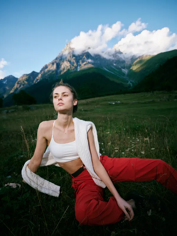 Outdoor scene of a woman in a white crop top and red pants relaxing on a grassy meadow with rugged mountains and a blue sky, soft sunlight and serene alpine scenery