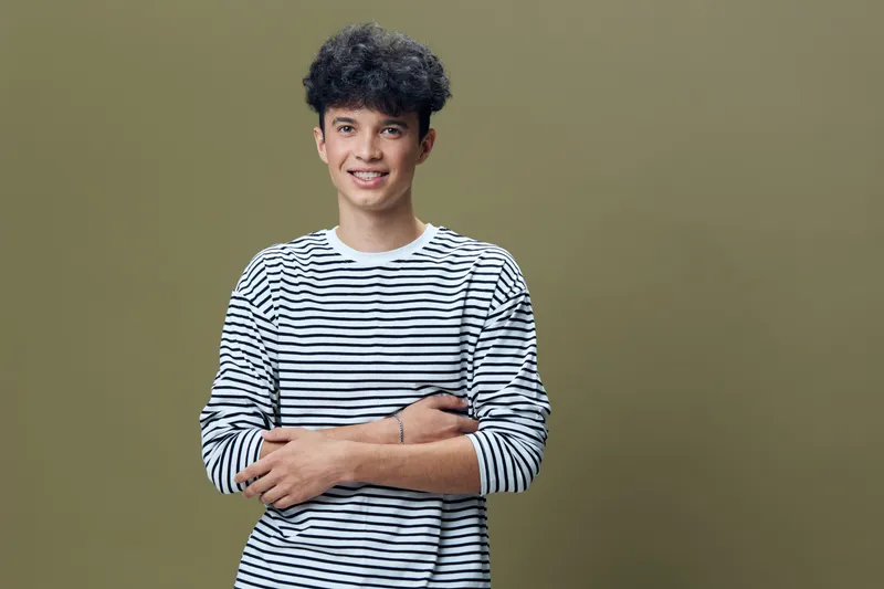 Smiling young man in striped shirt with curly hair standing confidently against a muted olive background, expressing positivity and casual style in a relaxed pose.