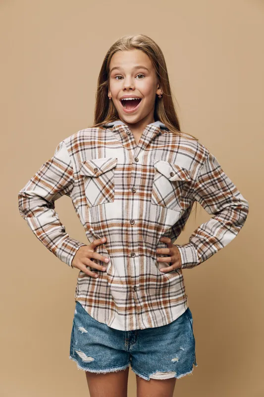 Girl with hands on hips wearing plaid shirt and denim shorts stands cheerfully smiling on beige background. Child showing confident playful expression in casual outfit indoors.
