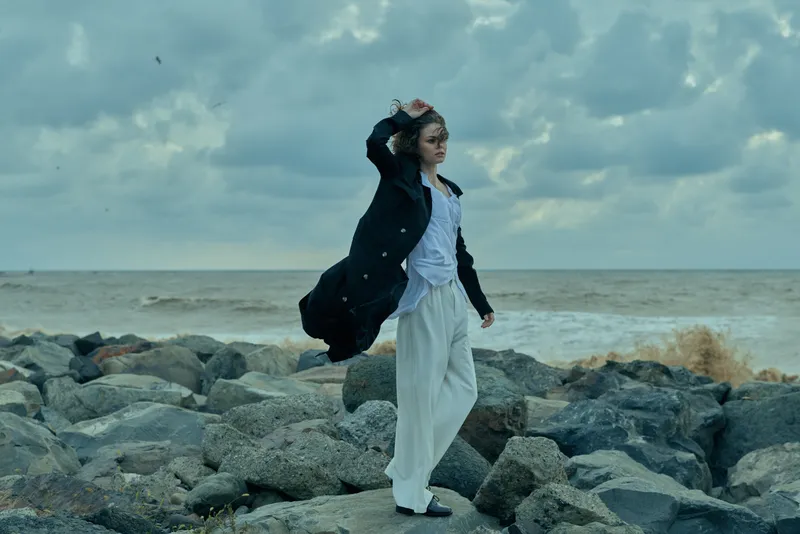 A woman stands on a rocky shoreline, her coat billowing in the wind, sea extending to the horizon, moody clouds overhead, a cinematic fashion moment captured outdoors on a coastal landscape