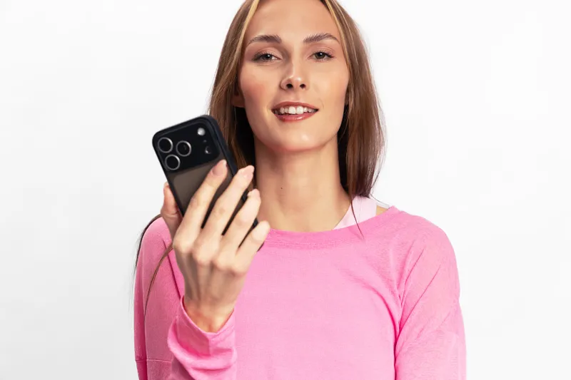 Young woman in pink sweater holding smartphone and smiling at camera isolated on white background. Concept of technology, communication, social media, and modern lifestyle.