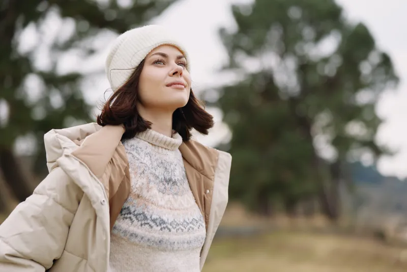 Woman in a light beige coat and knitted sweater stands outdoors, wearing a white beanie and gazing upward, surrounded by a blurred natural backdrop of trees and open fields on a chilly day.