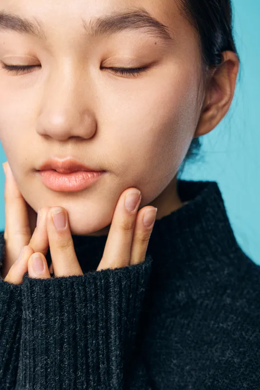Woman with closed eyes touching face with fingers in cozy black knitted sweater on blue background. Relaxation, skincare and calmness with natural look and peaceful expression.