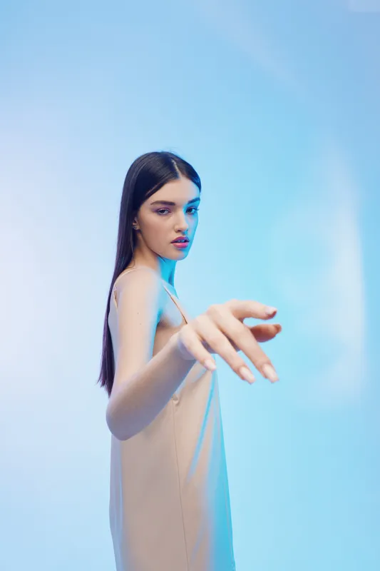 Young woman with long dark hair wearing a beige dress reaches out with her hand against a soft blue background. Concept for beauty, fashion, and elegance in studio lighting.