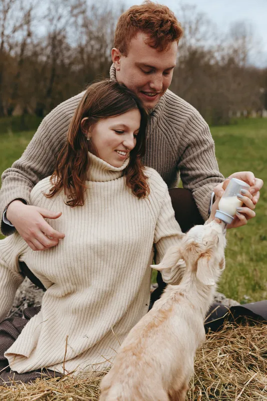 Warm couple outdoors feed a baby goat with a bottle, cozy sweaters, and a rustic field setting. A lifestyle moment about bonding, care, and farm life.