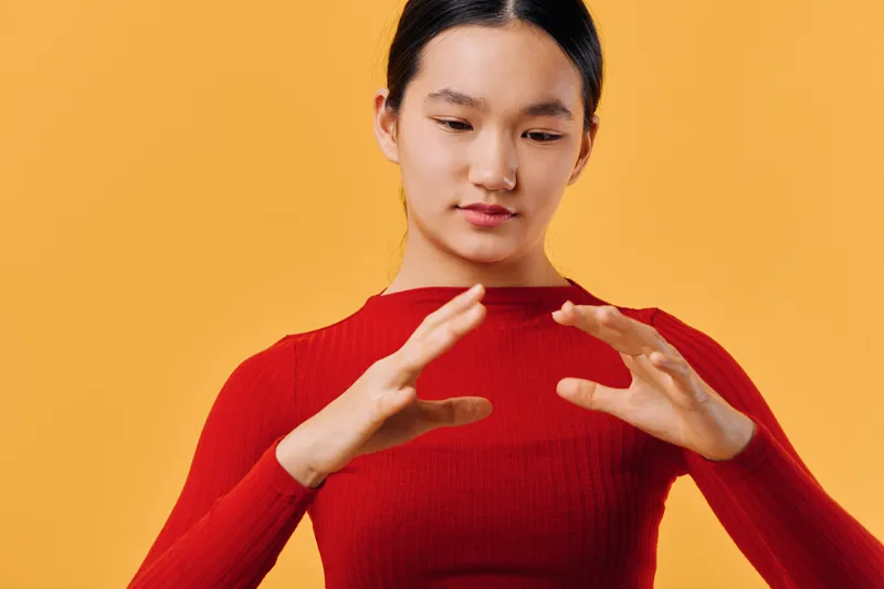 Young woman in red sweater on orange background concentrating and expressing with hands in gesture for communication or meditation, studio portrait with thoughtful mood.