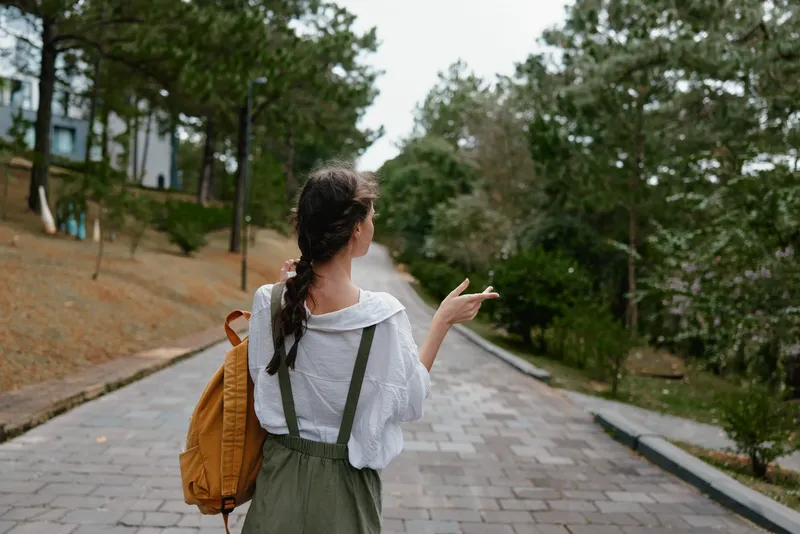 Young woman in casual outfit walking outdoors, enjoying nature in a park, with a serene expression and a backpack, surrounded by greenery in a tranquil setting