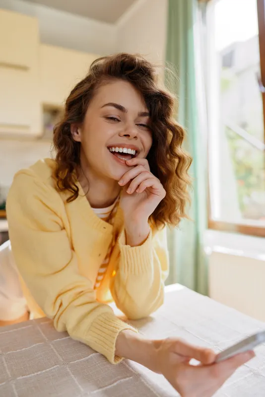 Bright indoor moment shows a young brunette woman in a yellow sweater smiling while texting on a phone at home, seated at a desk with natural light, her relaxed posture conveying a casual friendly