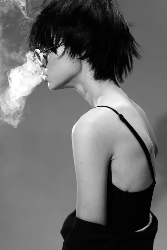 Black and white portrait of a woman exhaling smoke with short hair and glasses, stylish and moody atmosphere.