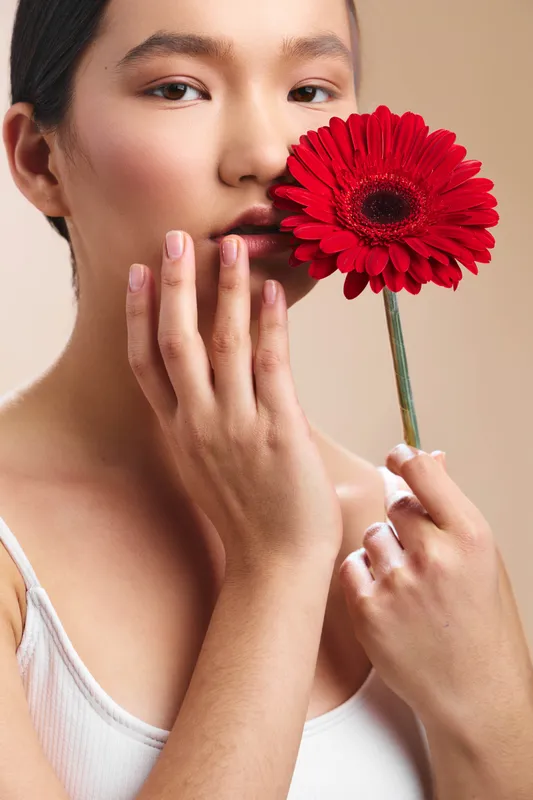 asian woman holds a vivid red flower close to her lips, expressing subtle grace and calm. the close up conveys beauty, softness, and a serene studio mood.