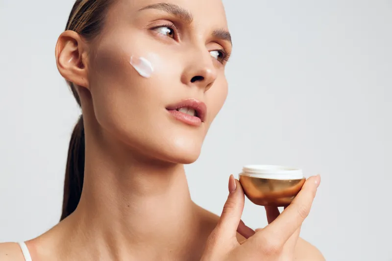 A young woman applies face cream on her cheek holding a jar in her hand. The studio background is plain white, highlighting skincare and beauty concepts.