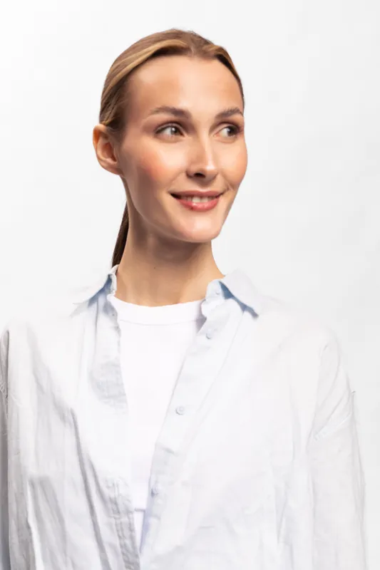 Young woman with light brown hair wearing white shirt smiles gently and looks to the side. Bright studio portrait with soft lighting on white background. Concept of natural beauty and positivity.