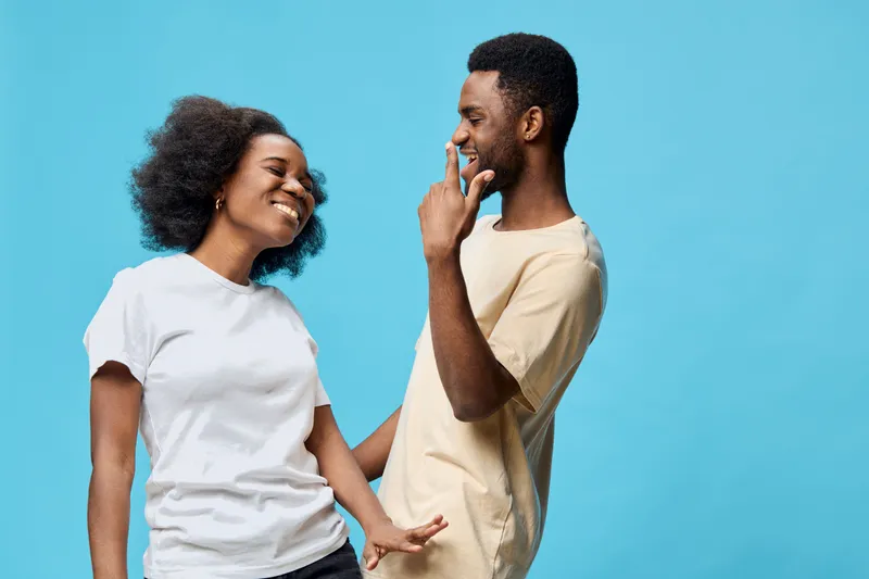 A joyful couple dressed casually in white and beige shirts, interacting playfully against a bright blue background, reflecting love and happiness in a vibrant, youthful style