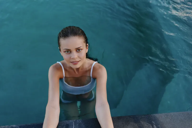 Cheerful woman in a pool reaching for the edge, sunlight on skin, clear water around, relaxed smile, summer lifestyle moment, casual swimwear, serene atmosphere, natural tones